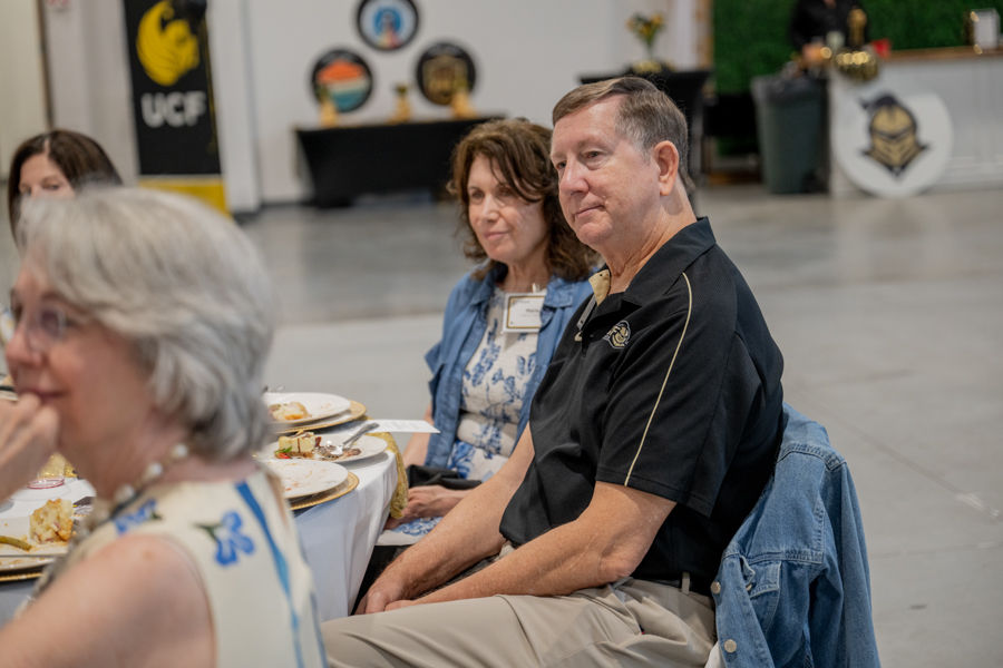 People seated at a round table during a social event, listening attentively to something off-camera and eating dessert plates nearby and wearing name tags.