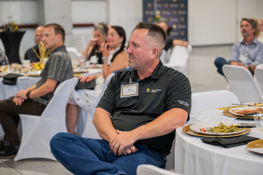 Man wearing a name badge sits at a round banquet table during a conference, listening to a speaker.