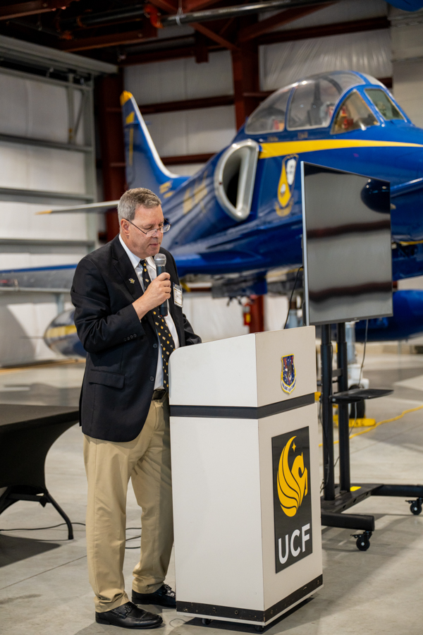 Man in a blazer giving a speech at a podium in an aircraft hangar, with a blue and yellow aircraft behind and a UCF logo on the lectern.