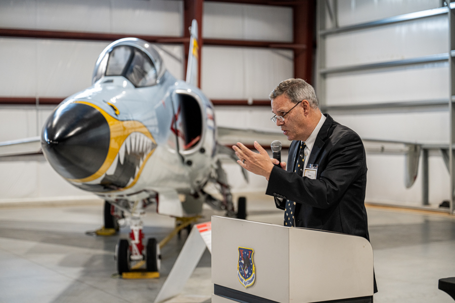 Man in a suit at a lectern, speaking into a microphone beside a fighter jet in a hangar.