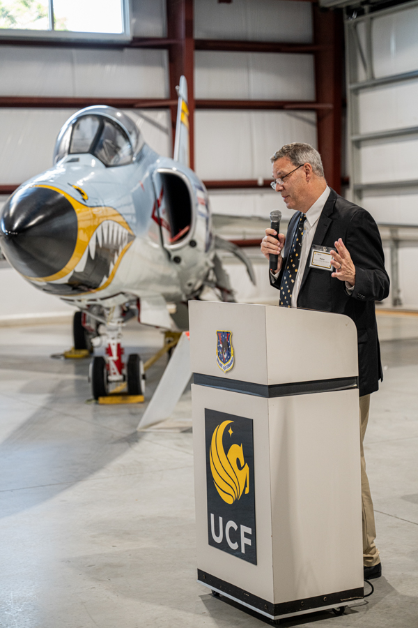 Man in a suit giving a speech at a podium labeled UCF, in a hangar beside a silver fighter jet.