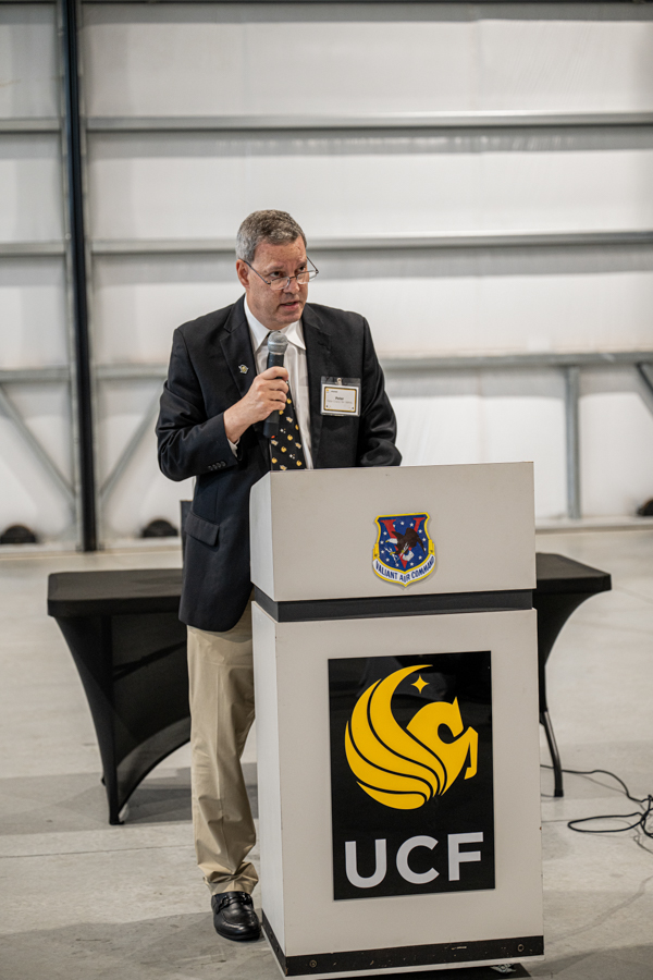Man in a blazer and tie speaks into a microphone at a podium with UCF logo in a large indoor space.