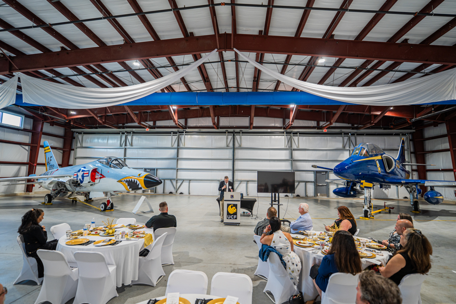 Formal dinner inside an aviation hangar with two vintage aircraft flanking the room; guests sit at round tables while a speaker stands at a podium in the center.
