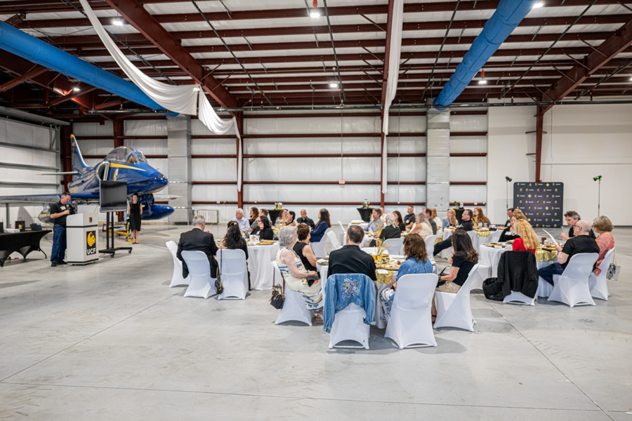 Formal luncheon in a hangar with a blue airplane on the left; guests sit at round tables with white chair covers.