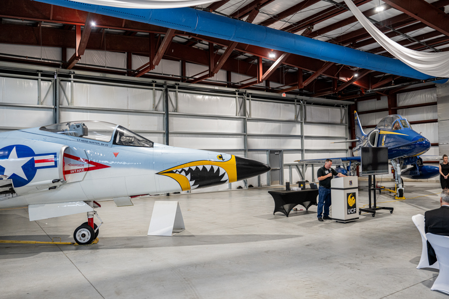 Light-blue fighter jet with shark-mouth nose art in a hangar, audience seated, speaker at a podium with another blue plane in the background.