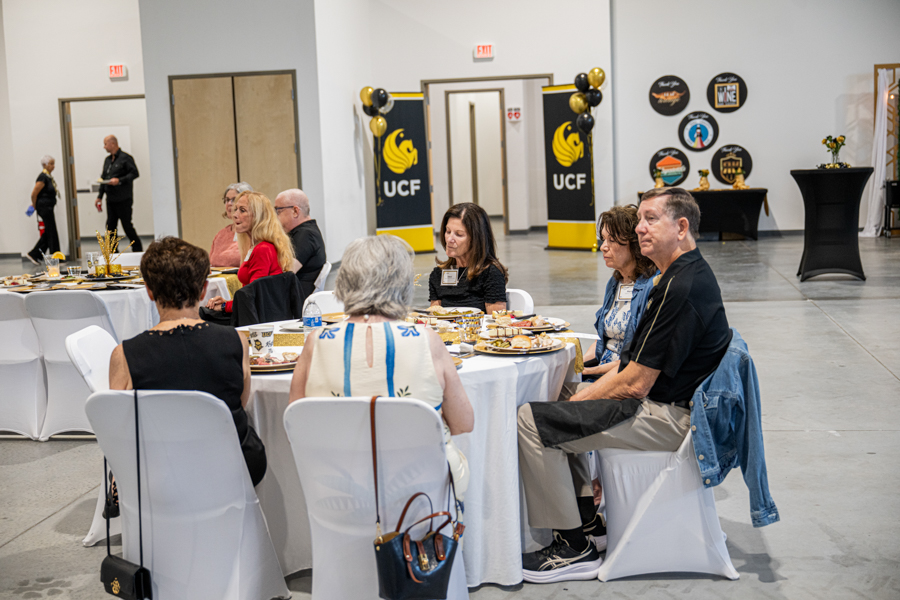Group of adults seated around a round table with plates of food at an indoor reception; UCF banners are visible in the background.