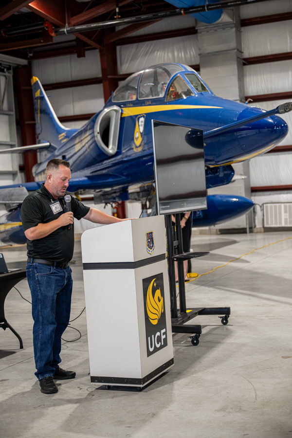 Man in a black shirt speaks into a microphone at a podium with a UCF crest, in an aviation hangar beside a blue jet.