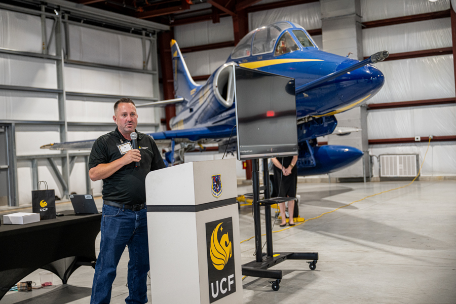 Man speaks into a microphone at a podium in a hangar, a blue jet in the background with UCF branding nearby.