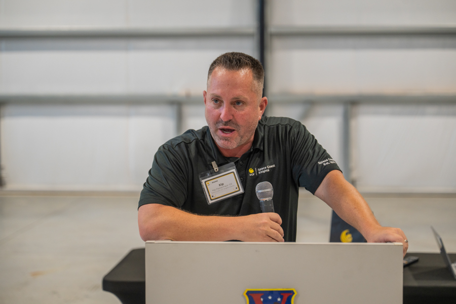 Male speaker at a lectern, speaking into a handheld microphone, wearing a name badge in a large indoor space.