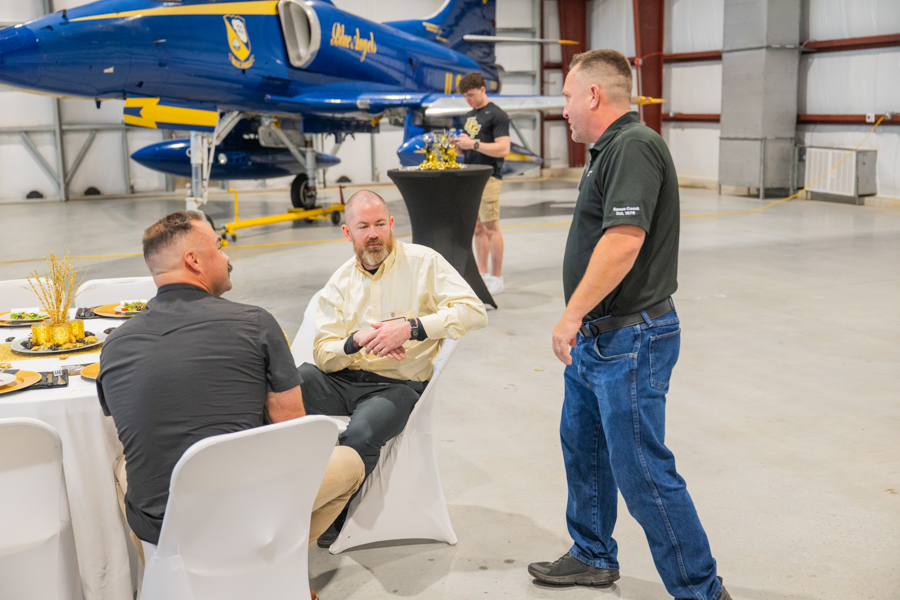 Three men sit at a round table with white linens in an aircraft hangar, a blue jet in the background; one man stands and talks nearby.