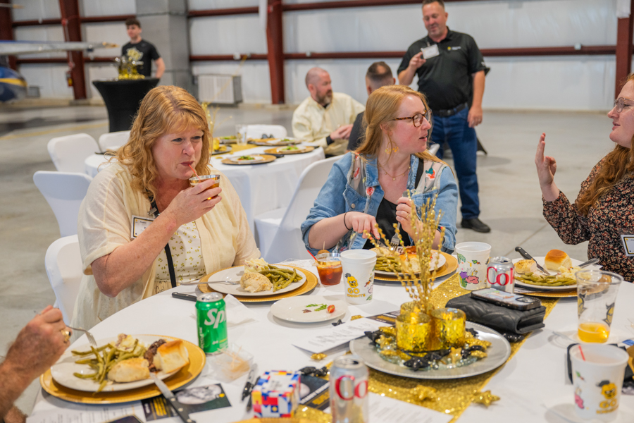Group of people seated at a round banquet table in an industrial-style hall, eating and chatting during a catered event.