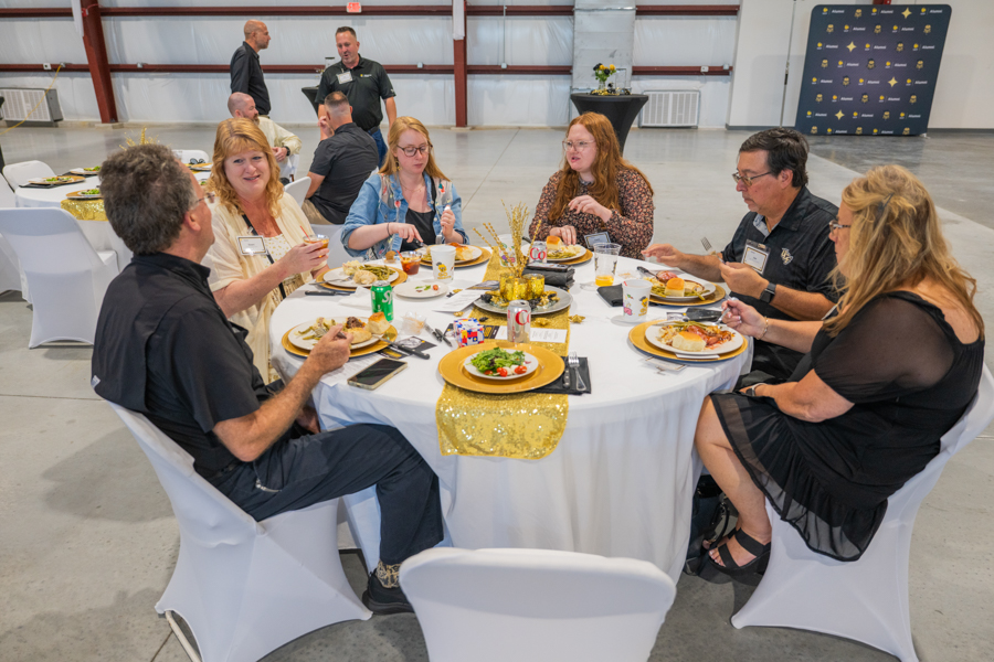 Seven adults sit around a round banquet table in a large venue, sharing a meal and conversation with plates, drinks, and a gold centerpiece.