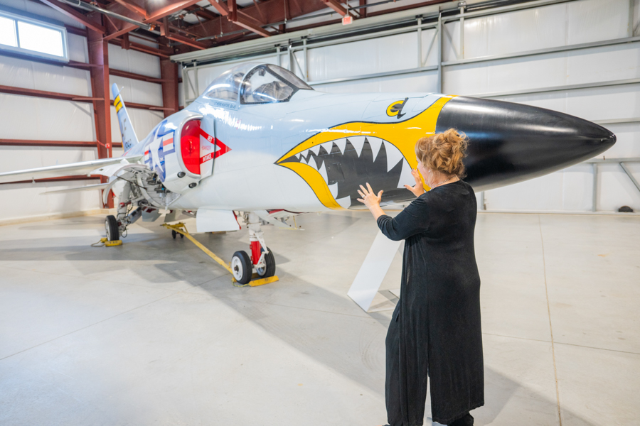 Person in a black coat touching a fighter jet with shark-mouth nose art inside a hangar at a museum or maintenance facility.