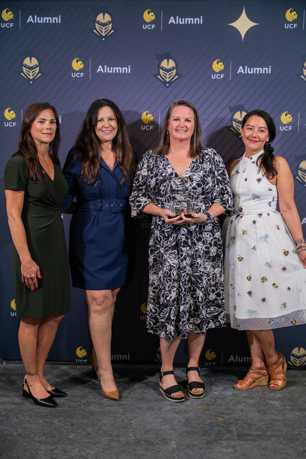 Four women stand together at a UCF Alumni event backdrop; the center woman holds an award.