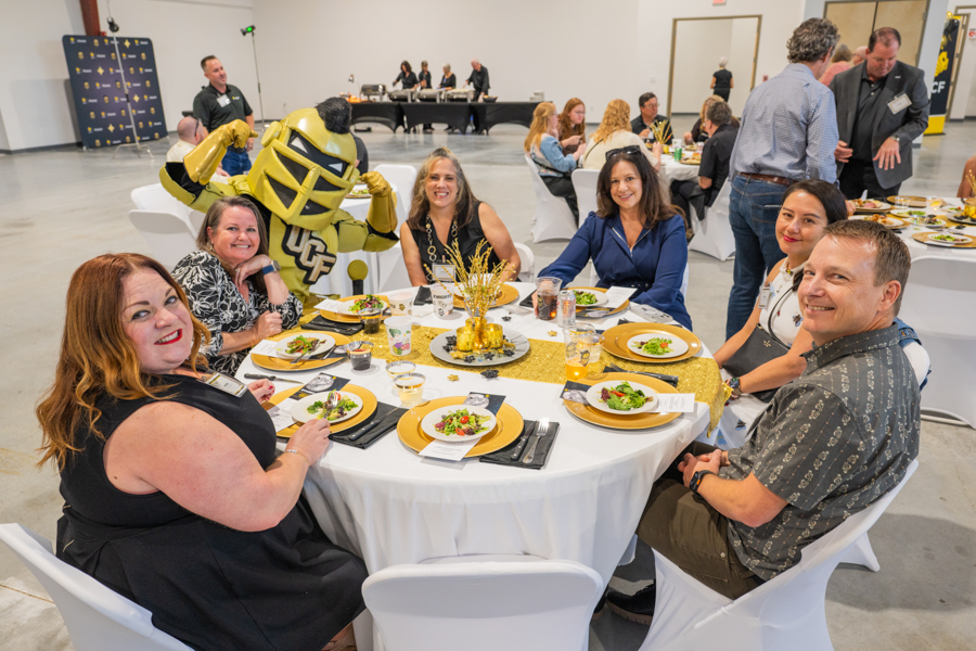 Eight people sit around a round table at a formal event, smiling for the camera; a yellow-and-black mascot stands behind them with raised arms. The table has gold placemats, a centerpiece, and plates of salad with drinks visible in front of guests.]