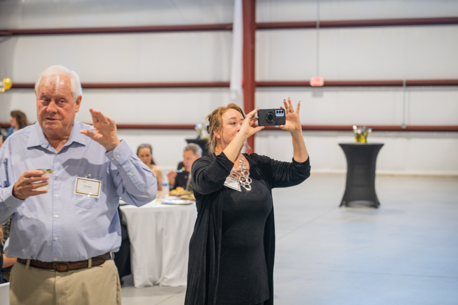 Woman in a black outfit takes a photo with a small camera at a social event; an older man with a name badge and a drink stands nearby.