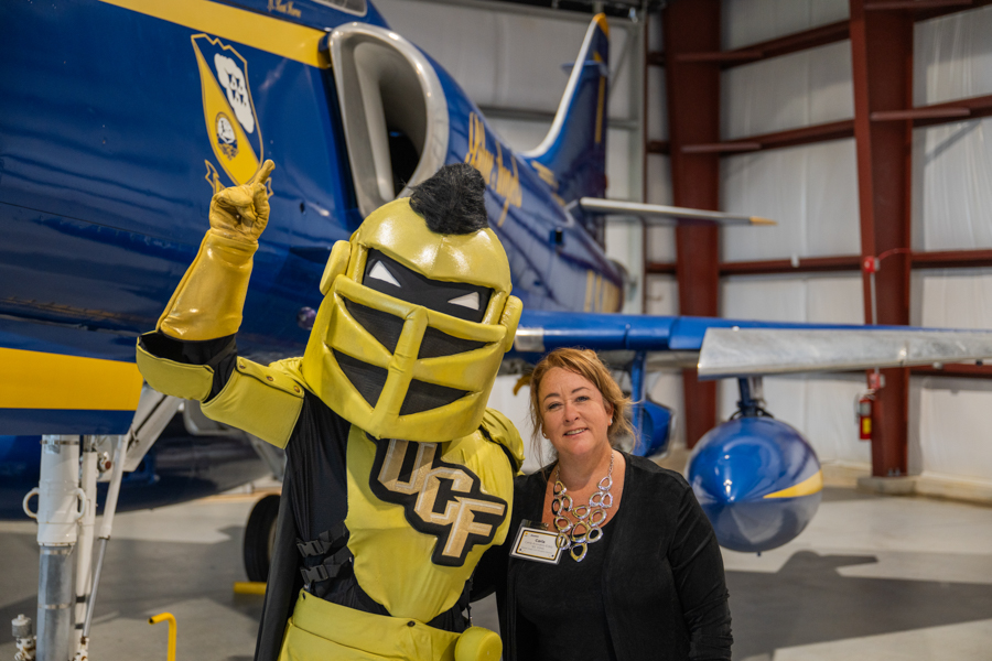 Costumed yellow armored mascot raises a finger beside a smiling woman next to a blue and yellow airplane in a hangar.