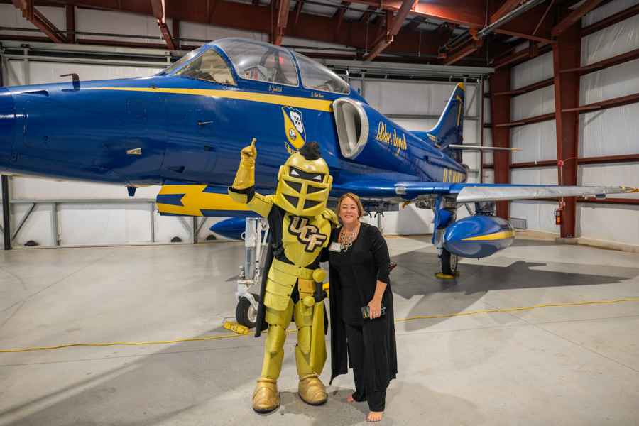 Yellow armored character with 'UCF' on chest poses with smiling woman in black, beside a blue and yellow jet in a hangar.