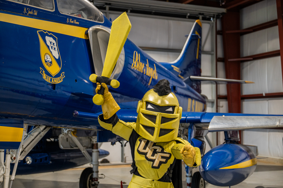 Yellow armored knight mascot holding a yellow sword in front of a Blue Angels jet in a hangar.