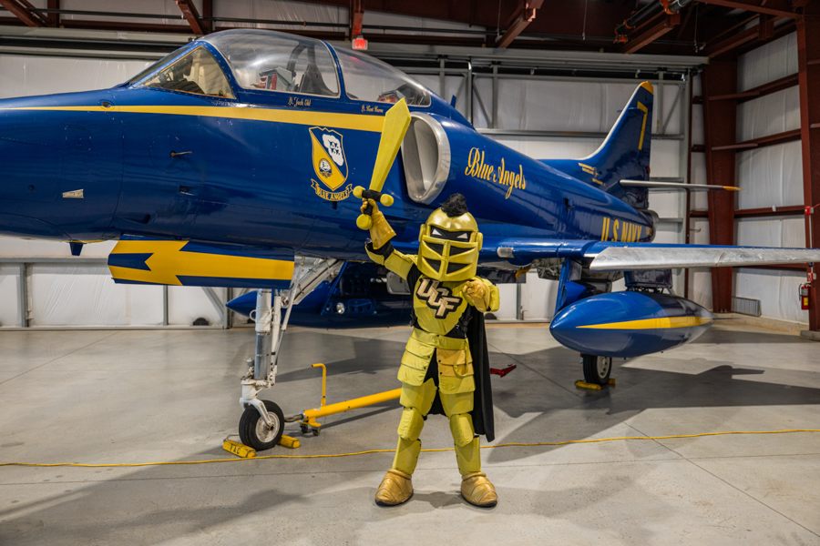 Blue Angels jet in a hangar with a person in bright yellow armor posing with a yellow sword in front of the plane's nose art.