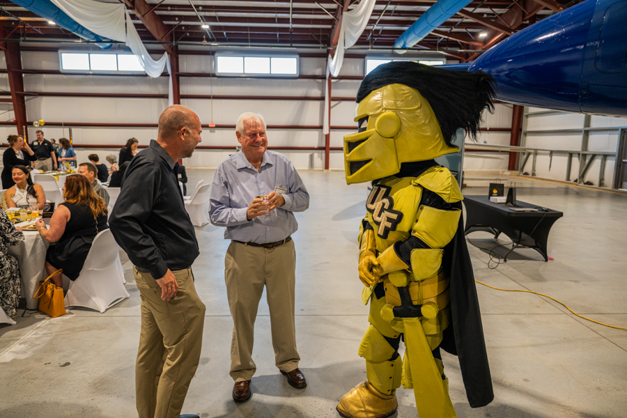 Two men chat with a yellow knight mascot in a large hangar, guests seated at tables in the background.