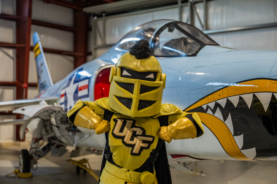 Yellow knight mascot with UCF logo poses in front of a vintage blue airplane with a shark-mouth design inside a hangar, arms outstretched.