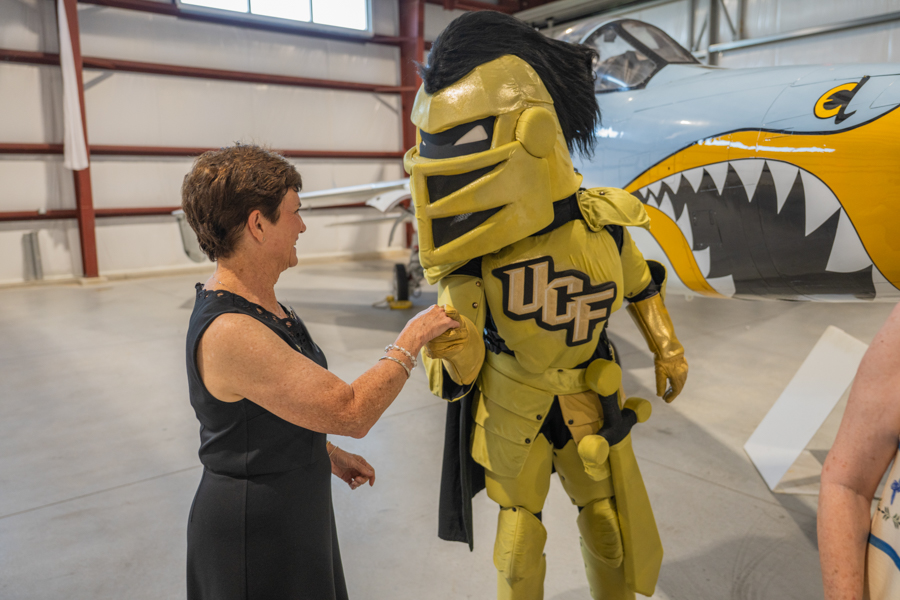 Woman in a black dress shakes hands with a gold UCF Knights mascot inside a hangar beside a plane with a shark-mouth design