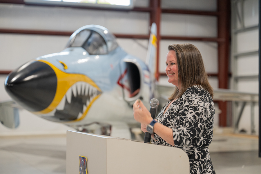 Woman in a black-and-white floral dress speaks into a microphone at a podium beside a shark-nosed jet inside an aircraft hangar.