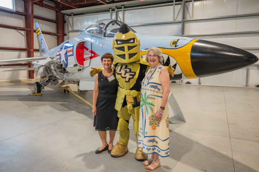 Two women pose with a yellow knight mascot beside a vintage fighter jet inside a hangar, smiling at the camera.