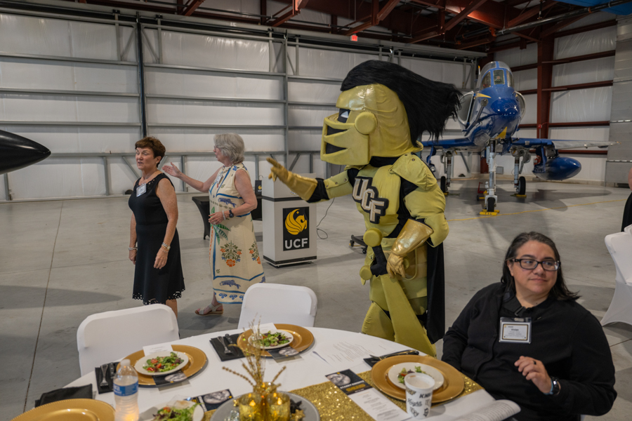 UCF Knights mascot in gold armor walks through a hangar beside guests at a banquet, with a blue helicopter in the background.