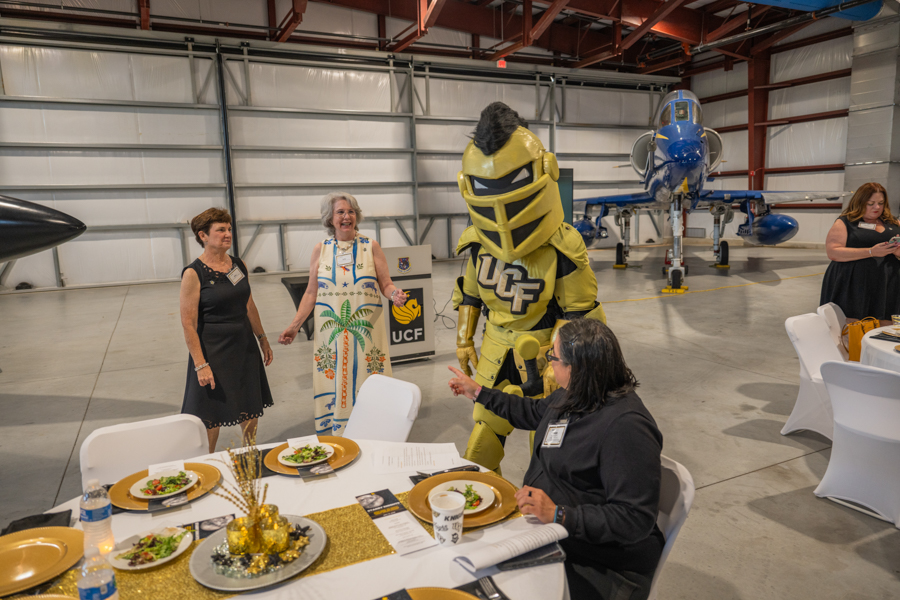 A yellow UCF mascot greets guests at a catered dinner in an aircraft hangar, with a blue jet visible in the background.