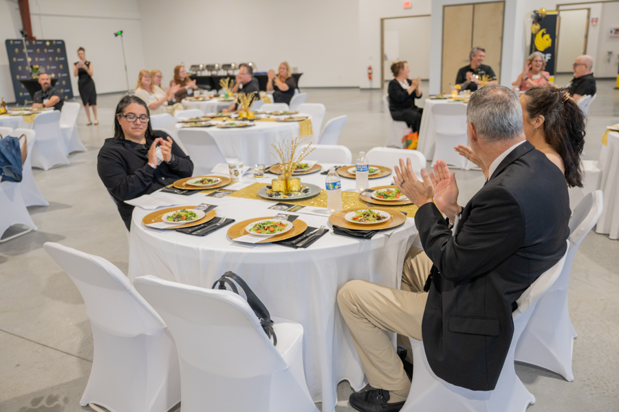 Guests seated at round tables with white tablecloths clapping during a formal dinner event in a large venue.