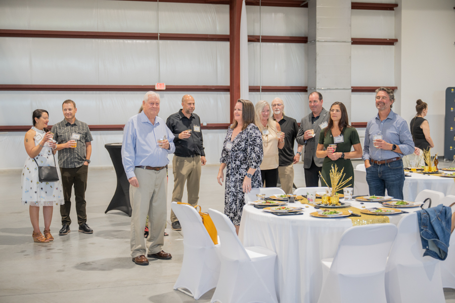 Group of professionally dressed adults mingling with drinks at a reception around round tables set for dining in a large venue.