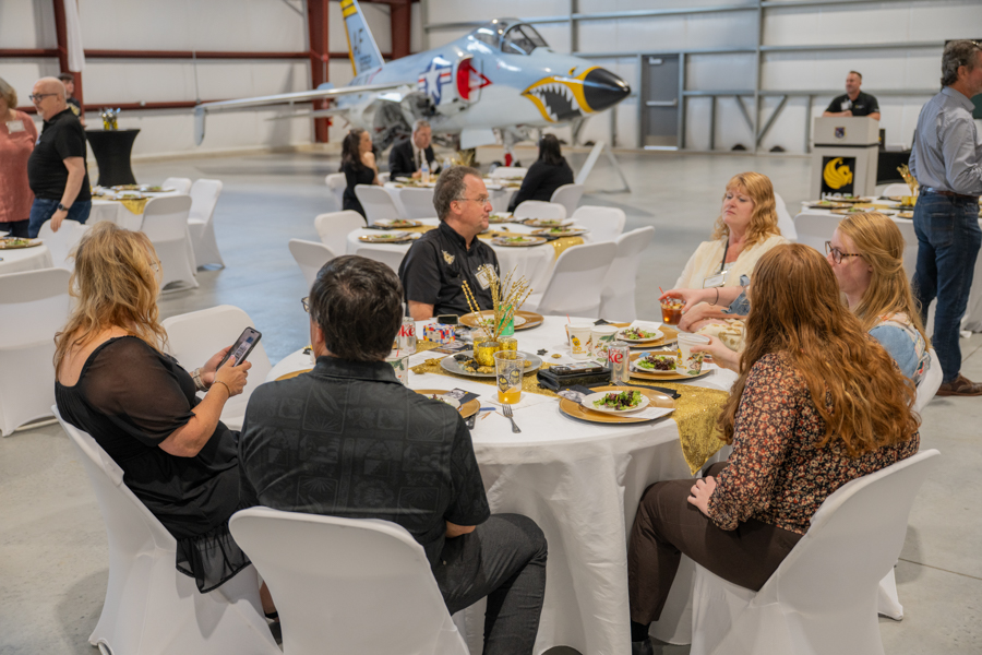 People seated around a round table in a hangar, eating and chatting, with a decorated jet in the background