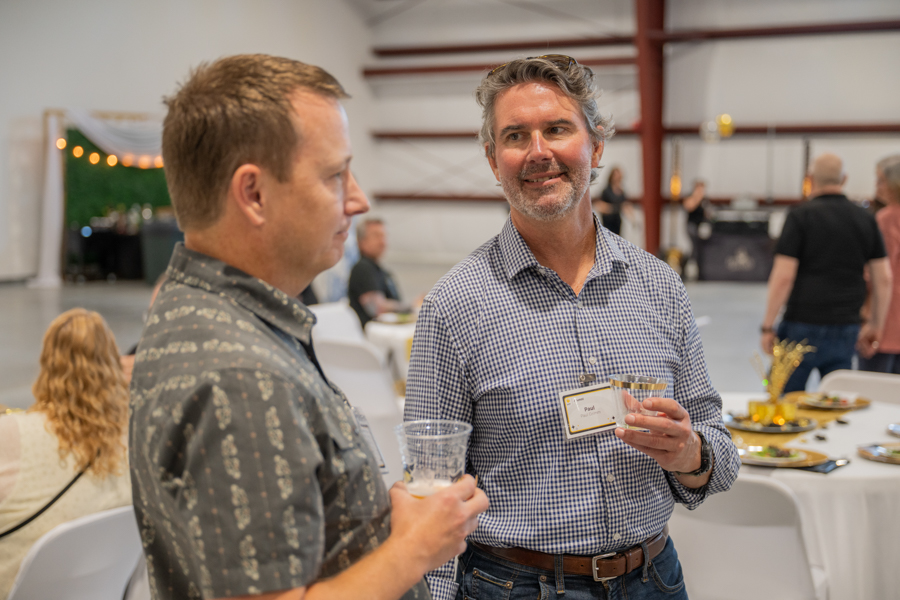 Two men chatting at a reception, each holding a cup; one wears a checkered shirt and a name badge in a large event space1 . (informative)
