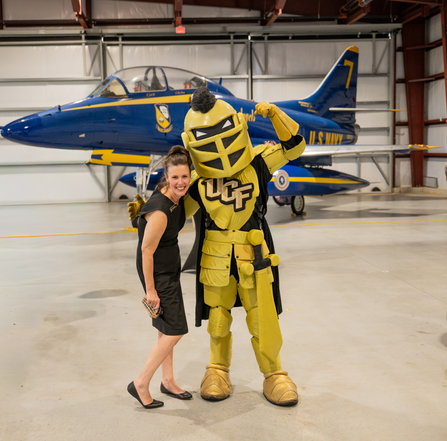 Smiling woman in a black dress posing with a yellow knight mascot inside a hangar, a blue Navy aircraft in the background.