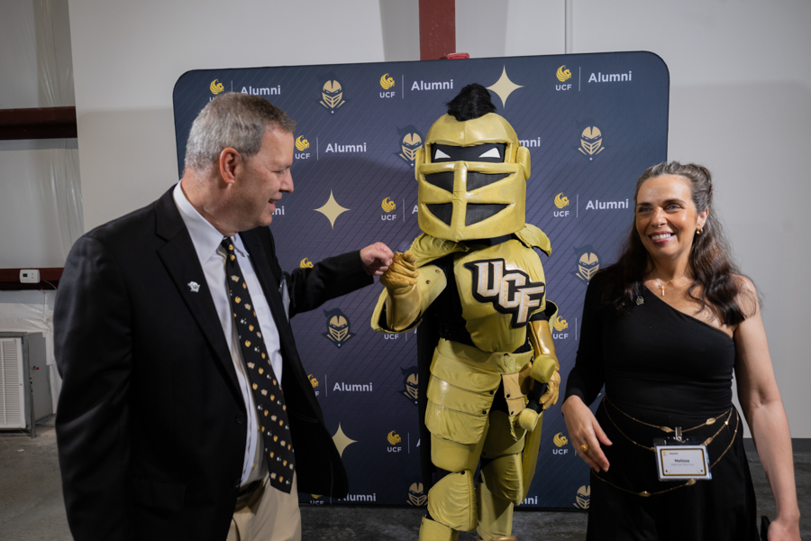 Two adults pose with a gold knight mascot in front of a UCF Alumni backdrop, smiling for the camera in an indoor event setting.