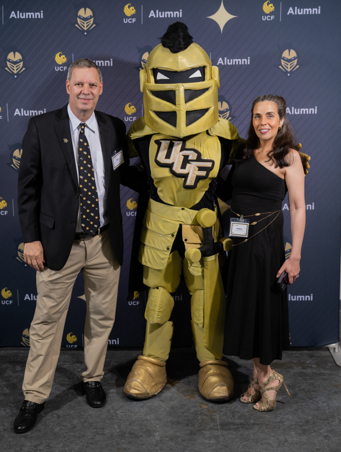 Man in a suit and a woman in a black dress pose with a large gold Knight mascot bearing the UCF logo at a UCF Alumni event backdrop.