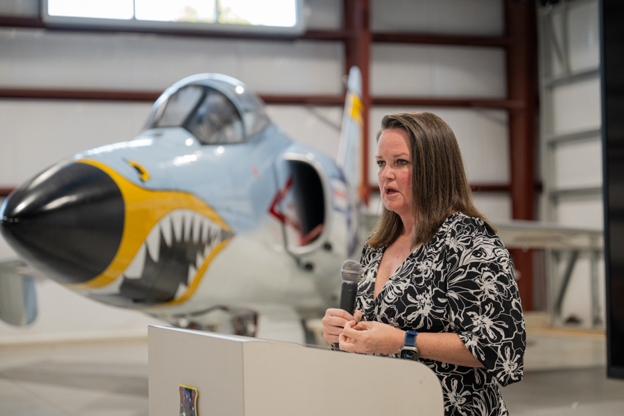 Woman speaking into a microphone at a podium in a hangar, with a vintage shark-mouthed fighter aircraft behind her.