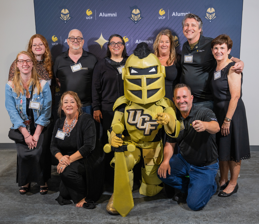 Group of people posing with a gold knight mascot in front of a UCF Alumni backdrop; everyone smiles at the photo.