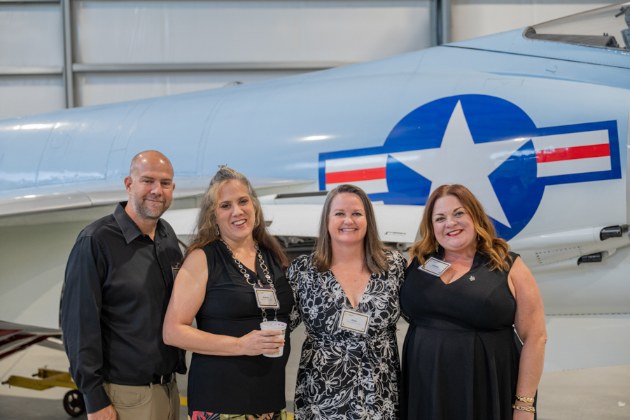 Four adults posing in front of a light-blue military jet with a white star insignia and red stripes inside a hangar.