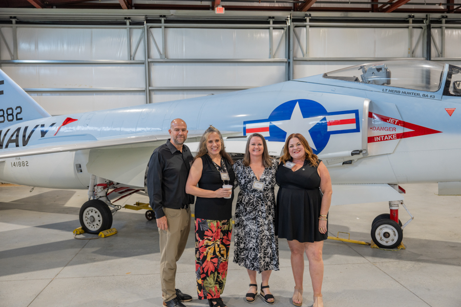 Group of four adults posing in front of a light-blue vintage military jet inside a hangar, smiling for the photo.