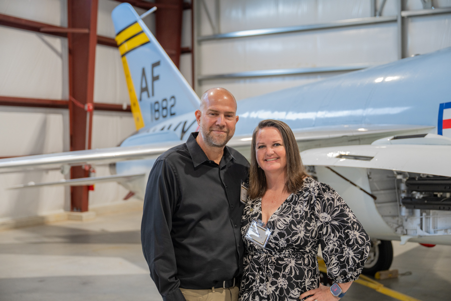 Smiling man and woman stand together in a hangar beside a blue military aircraft tail marked AF 1882.