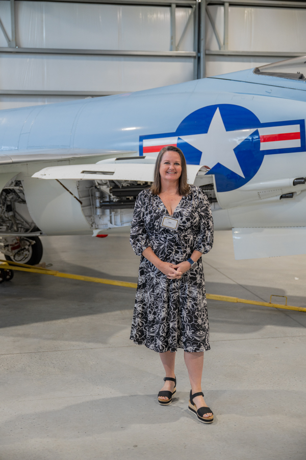 Woman in a black-and-white patterned dress stands in a hangar beside a silver military jet with a blue star insignia.