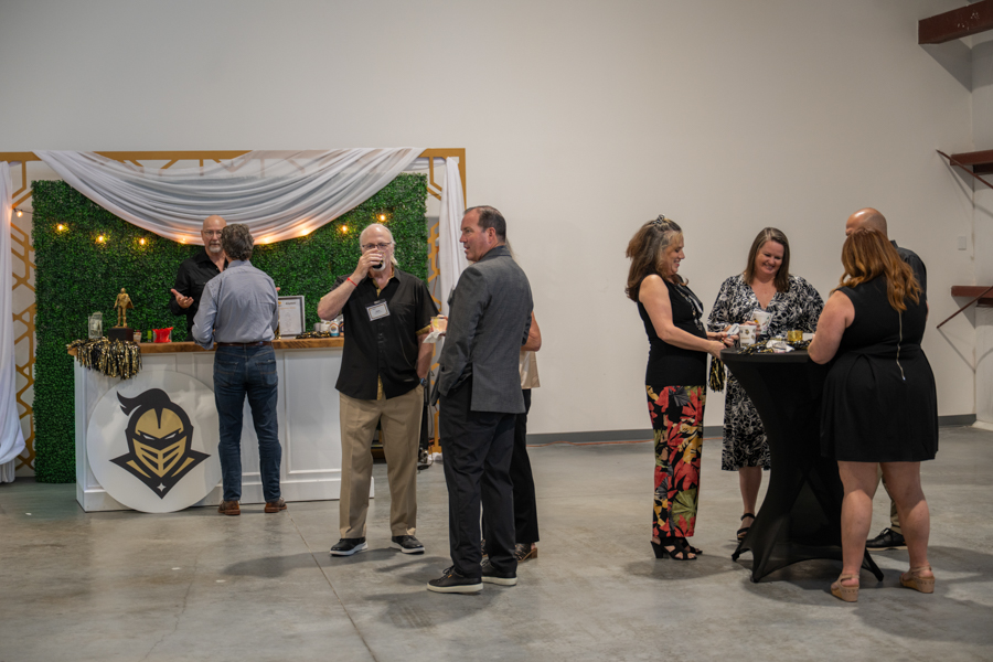 Guests mingle at a reception in a spacious venue; a decorated bar with greenery and white fabric backdrop in the background.