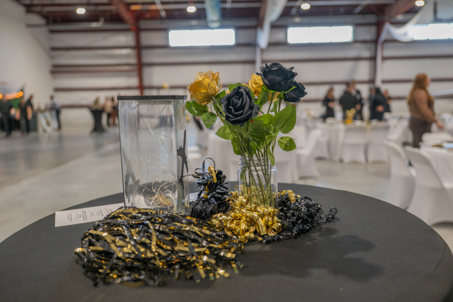 Table centerpiece with black and gold roses in a glass vase, surrounded by black and gold ribbons on a round table in a banquet hall