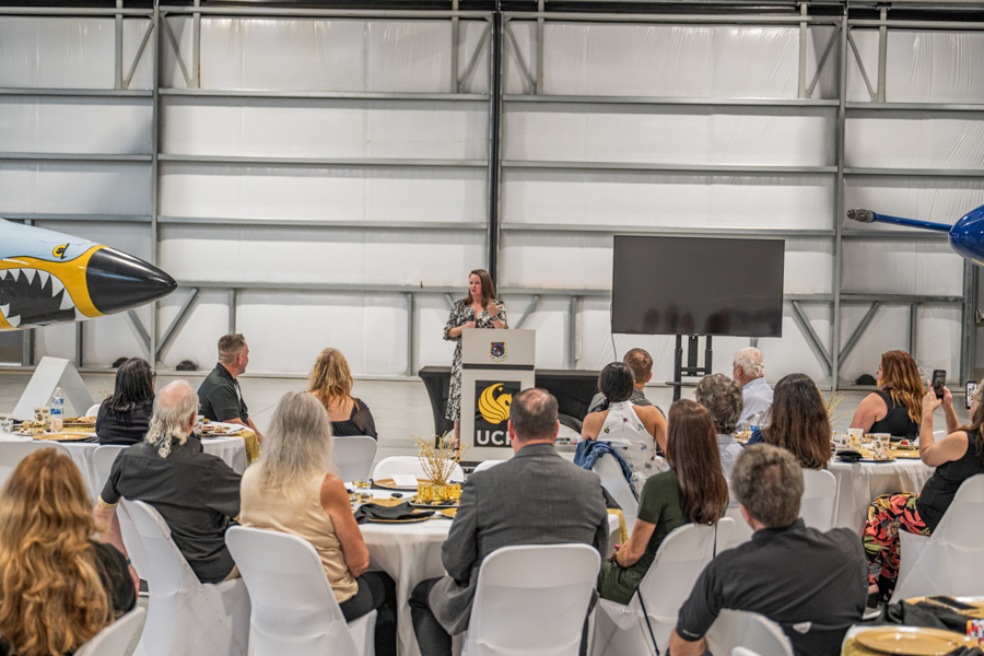Speaker at a podium addressing an indoor conference seated at round tables, with airplanes on either side in a hangar setting