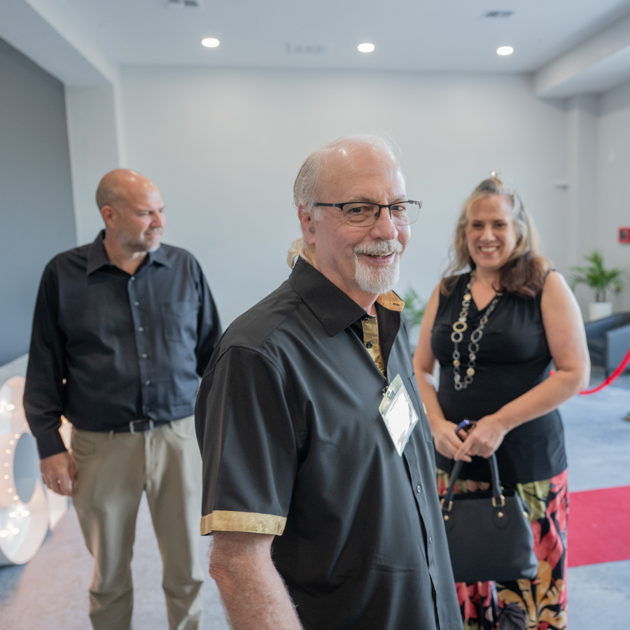 Smiling older man in a black shirt with a name badge in the foreground at an indoor social event, with two guests in the background.