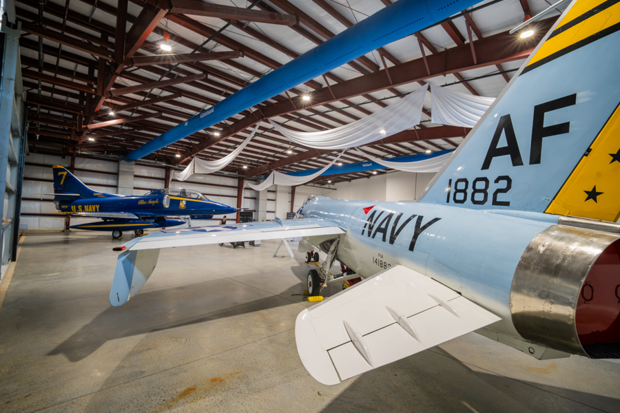 Inside a museum hangar showing a light-blue Navy fighter in the foreground with 'NAVY' on the fuselage and 'AF 1882' on the tail; a blue U.S. Navy jet is visible in the background amid white drapery and overhead beams.