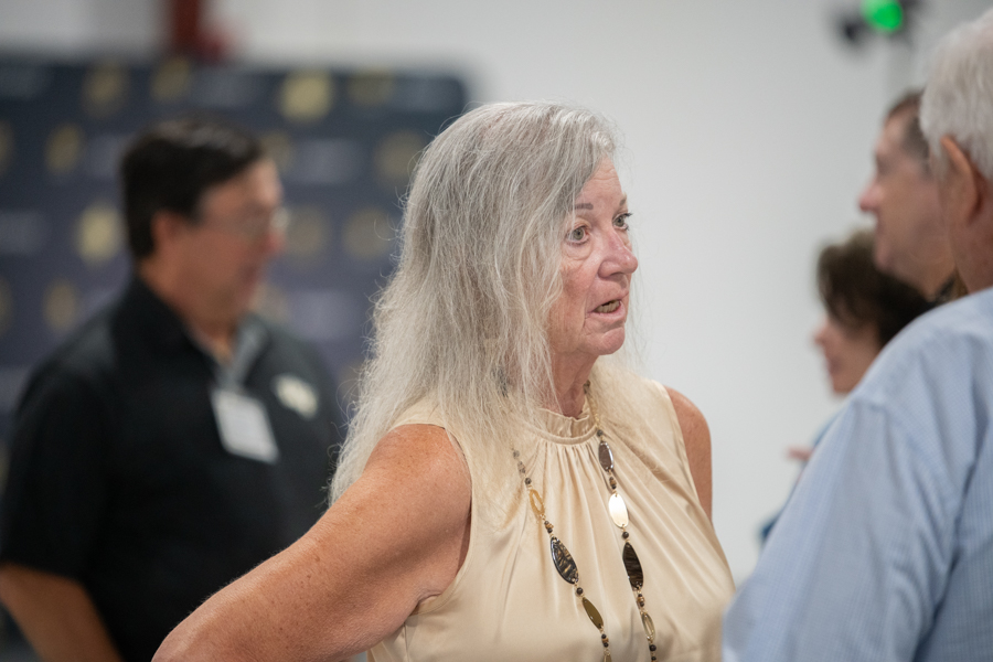 Older woman with long gray hair wearing a beige sleeveless top and decorative necklace talks to a man in a light blue shirt at a social event, with blurred background guests.